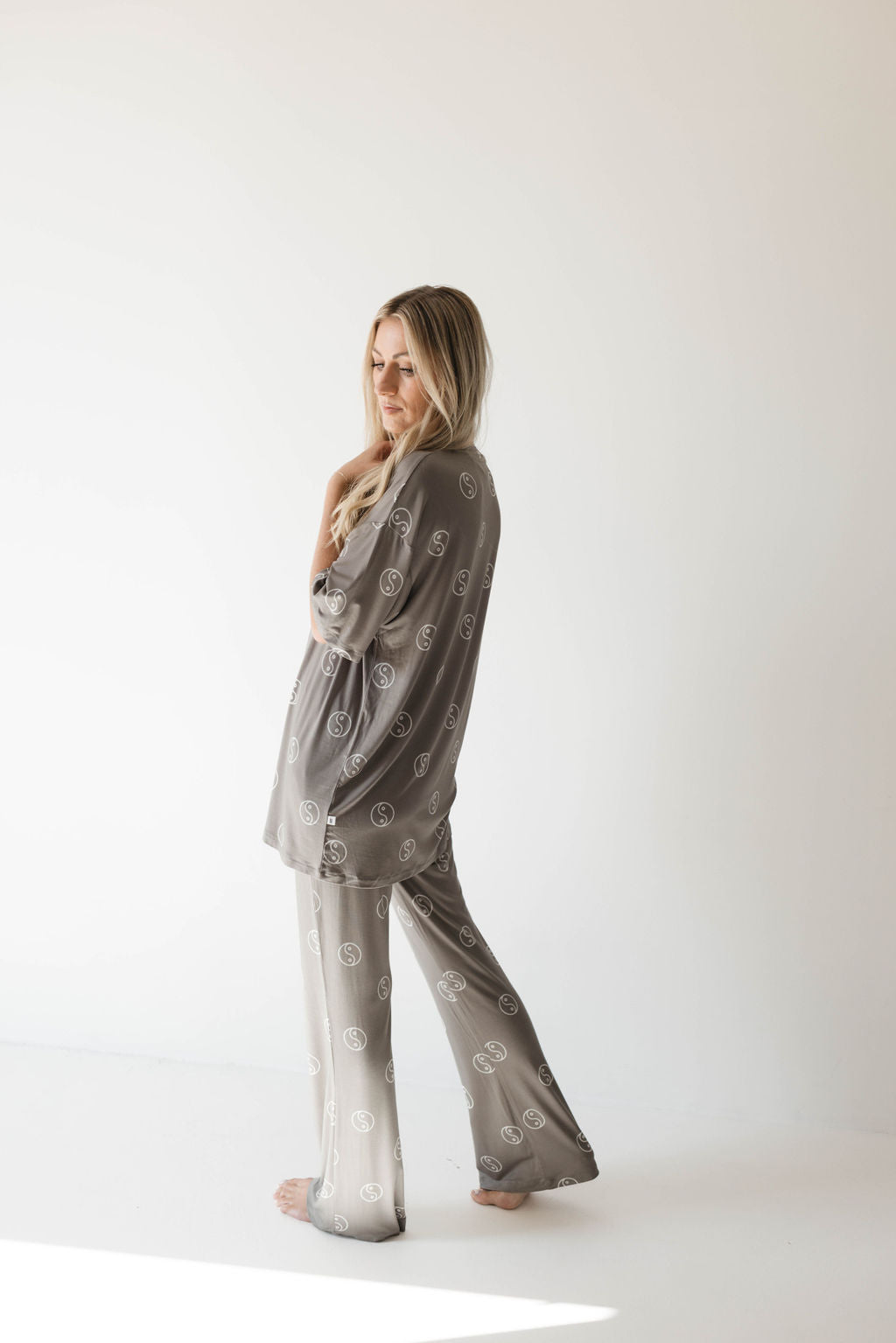 A woman with long blonde hair stands barefoot in a light, minimalistic space. She is wearing the Short Sleeve Women's Bamboo Pajamas in Sage Yin Yang from forever french baby, an oversized set made from breathable bamboo fabric adorned with a circular pattern. Her head is slightly tilted down, and she gently touches her face with one hand.