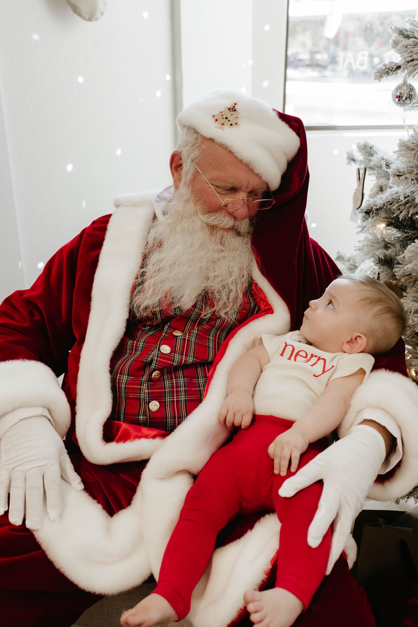 A man dressed as Santa Claus, complete with a white beard and a red and white suit, is holding a baby. The baby wears red pants and a “merry” shirt from Forever French Baby while gazing up at Santa. They stand next to a white-flocked Christmas tree—creating truly magical moments captured by Amanda Riley Photos in the Santa Photos | Forever French Baby x Amanda Riley Photos collection.