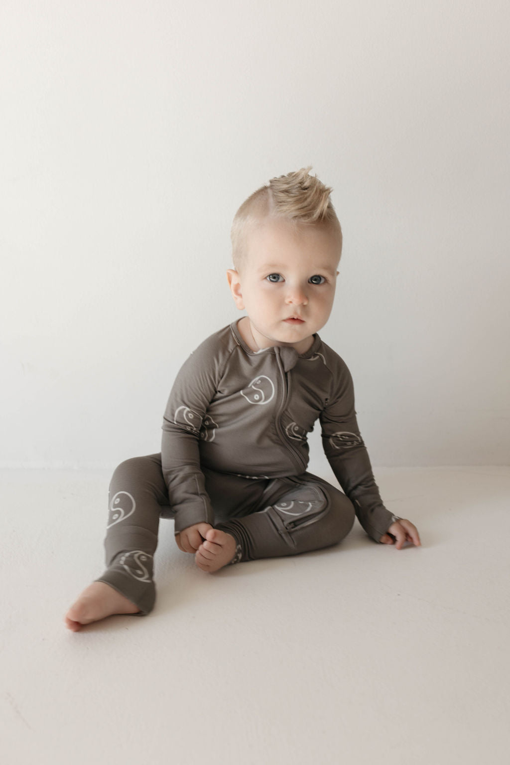 A baby with blonde hair sits on the floor, dressed in the Bamboo Zip Pajamas | Sage Yin Yang by forever french baby, featuring a matching gray set with a white swirl pattern. The baby gazes into the camera with a neutral expression against a plain, light-colored wall.