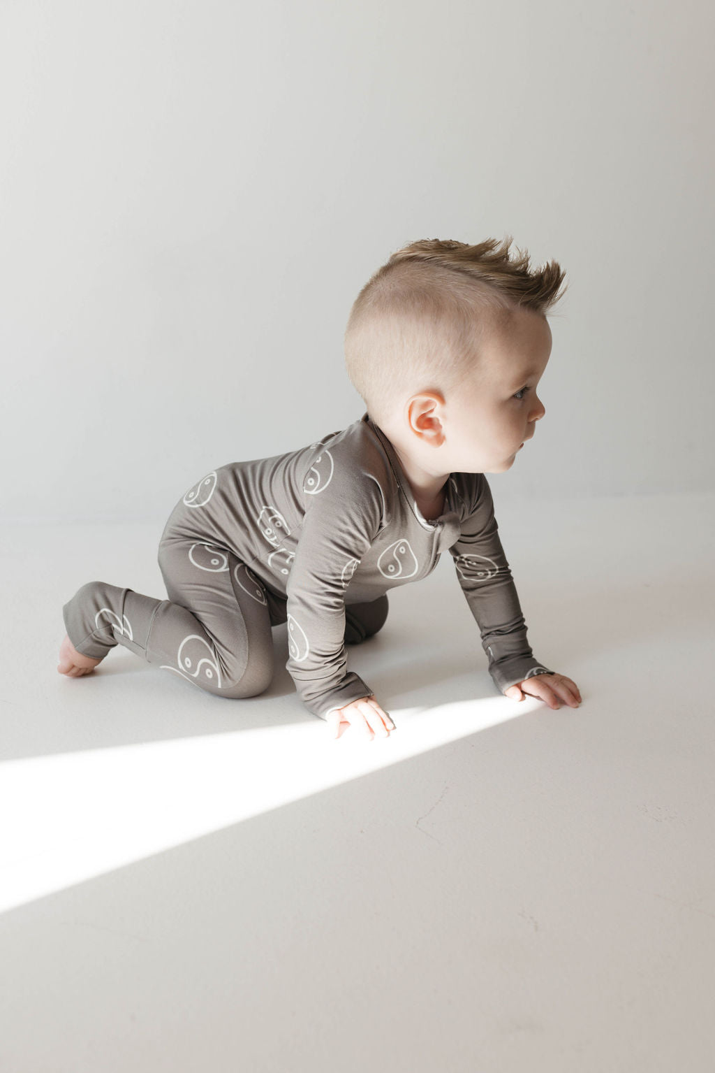 A baby is crawling on a white floor, dressed in breathable Bamboo Zip Pajamas from forever french baby. The Sage Yin Yang outfit features a subtle swirl design pattern and is made from hypo-allergenic fabric. The baby has short, light hair and is facing to the right, with light streaming in from the left side.