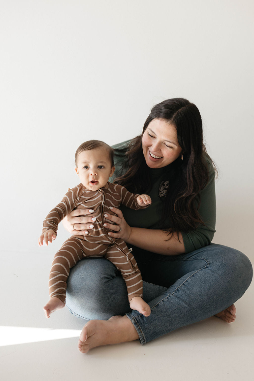 A woman with long dark hair, dressed in a green sweater and blue jeans, sits cross-legged on the floor. She cradles a baby who is looking at the camera and wearing hypo-allergenic Bamboo Zip Pajamas featuring a brown and white striped Static Waves design from forever french baby. Both mother and child appear happy and are smiling.