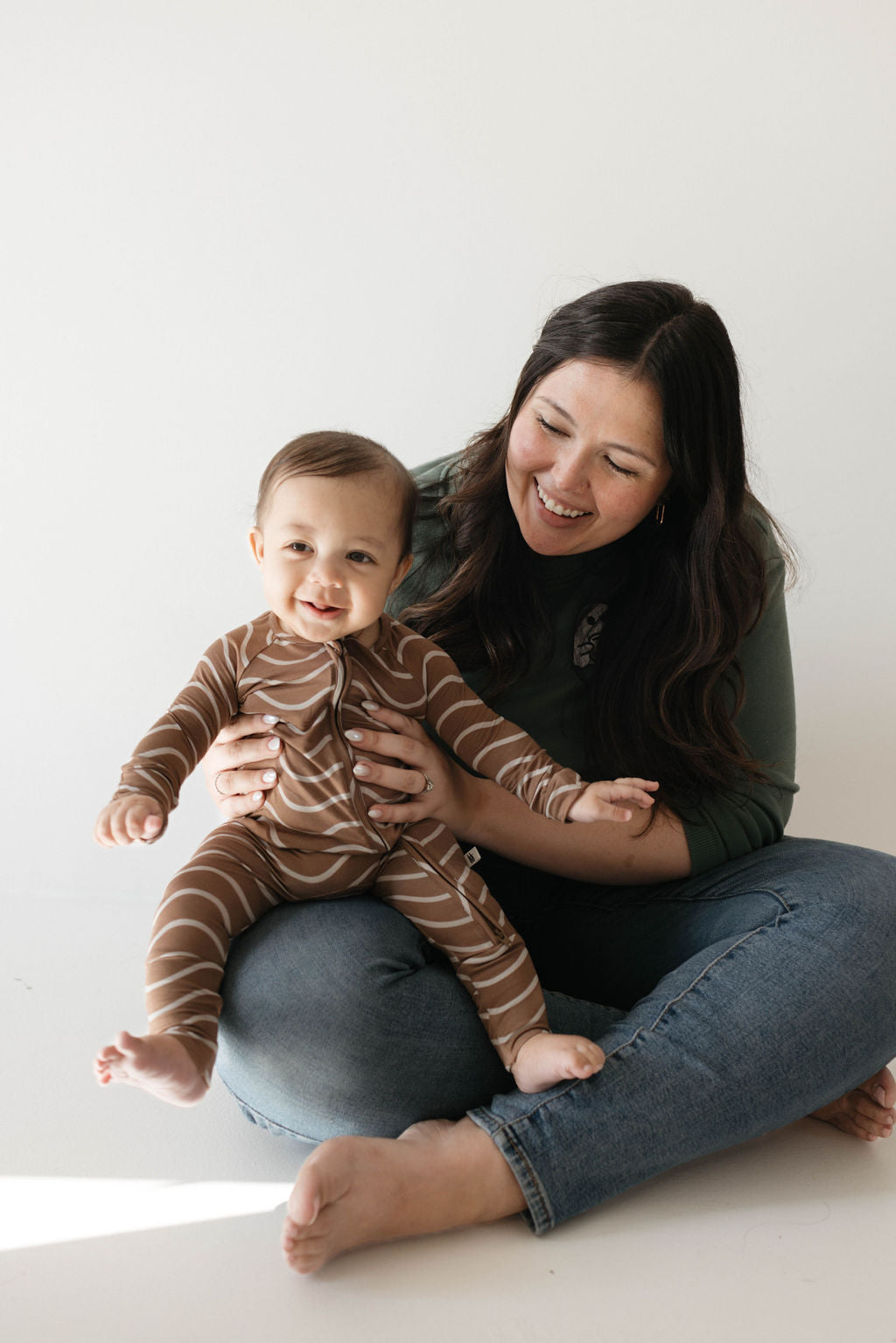 A woman with long, dark hair, dressed in a green sweater and blue jeans, sits cross-legged on the floor, smiling and holding a baby. The baby is wearing hypo-allergenic clothing—a brown and white striped onesie from the forever french baby brand's Bamboo Zip Pajamas collection called Static Waves—and is also smiling. Both appear to be enjoying a happy moment together.