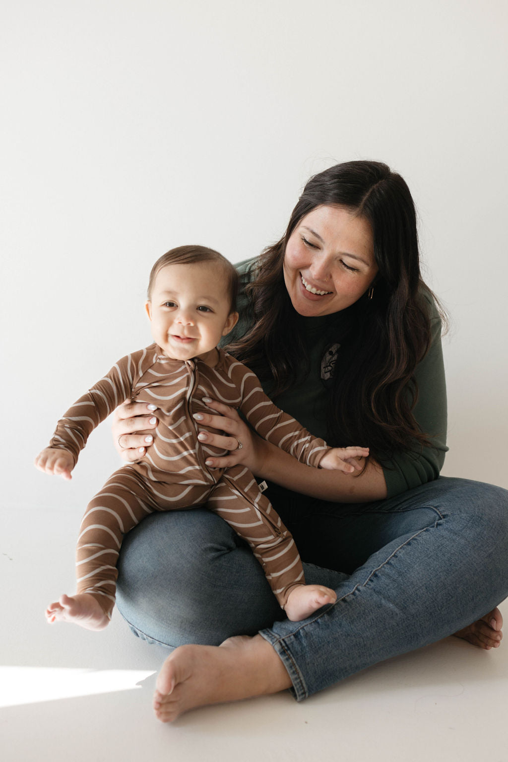 A smiling woman with long dark hair sits cross-legged on the floor, holding and supporting a smiling baby dressed in Bamboo Zip Pajamas | Static Waves by forever french baby. The background is plain white, creating a simple and cheerful atmosphere.