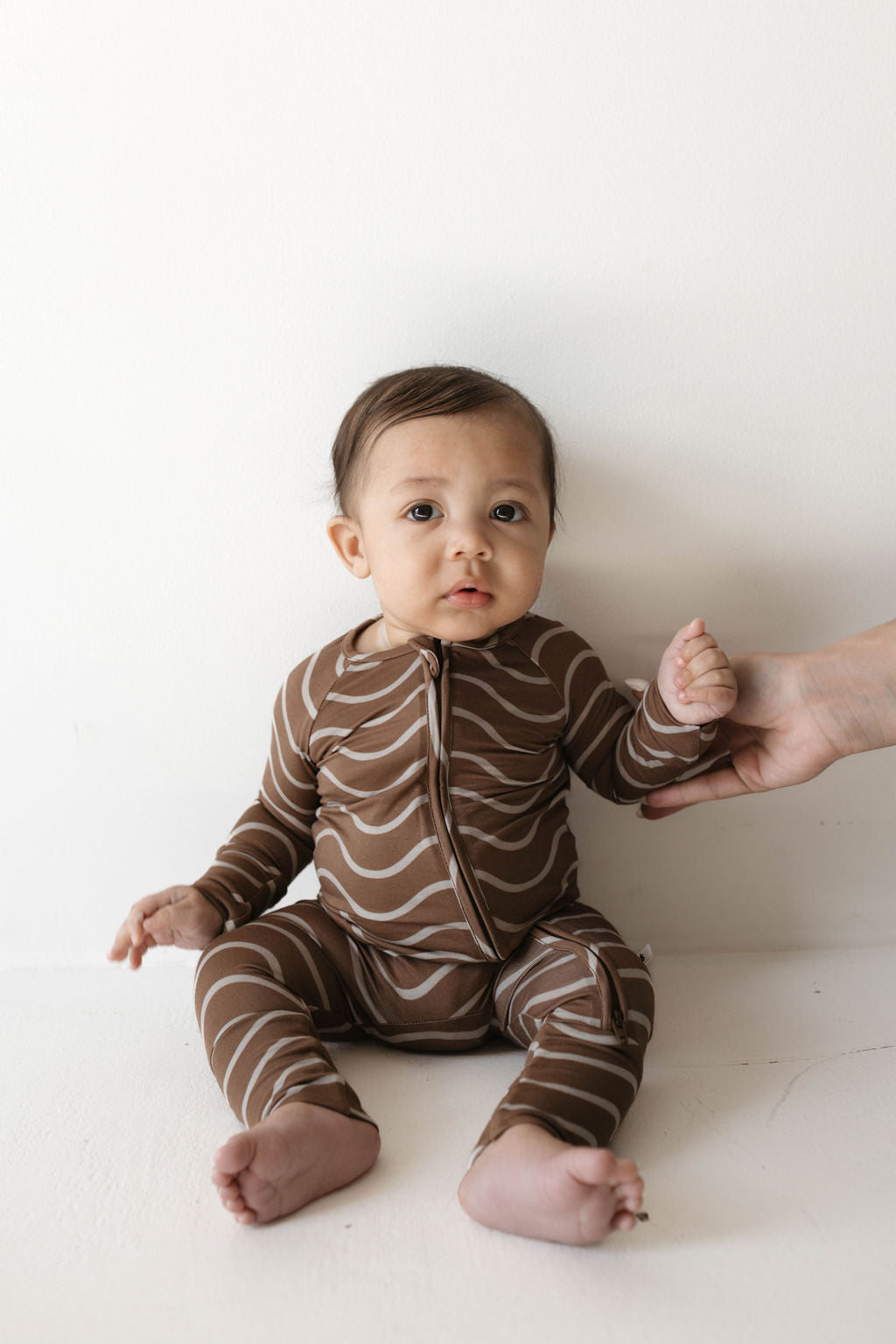 A baby in forever french baby's Bamboo Zip Pajamas | Static Waves sits on a white surface against a plain background. An adult hand gently supports the baby from the side. The baby, dressed in hypo-allergenic clothing, looks towards the camera with a curious expression.