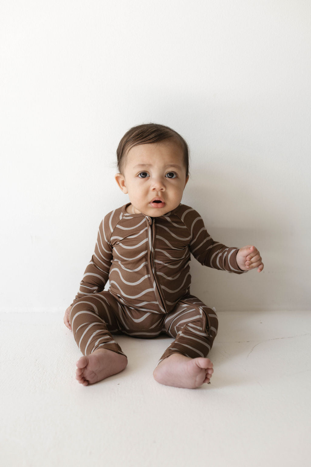 A baby sits on the floor against a plain white background, wearing Forever French Baby's adorable "Bamboo Zip Pajamas" in the Static Waves pattern. The baby has an inquisitive expression, looking slightly upward. Barefoot, with one hand resting on the floor for support.