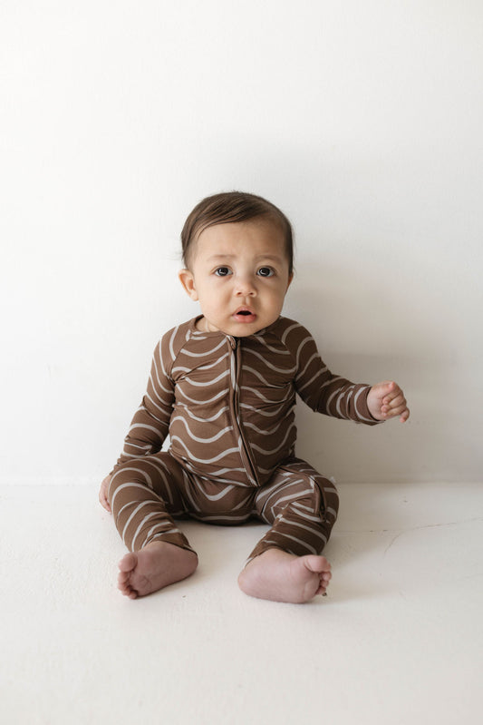 A baby sits on the floor against a plain white background, wearing Forever French Baby's adorable "Bamboo Zip Pajamas" in the Static Waves pattern. The baby has an inquisitive expression, looking slightly upward. Barefoot, with one hand resting on the floor for support.