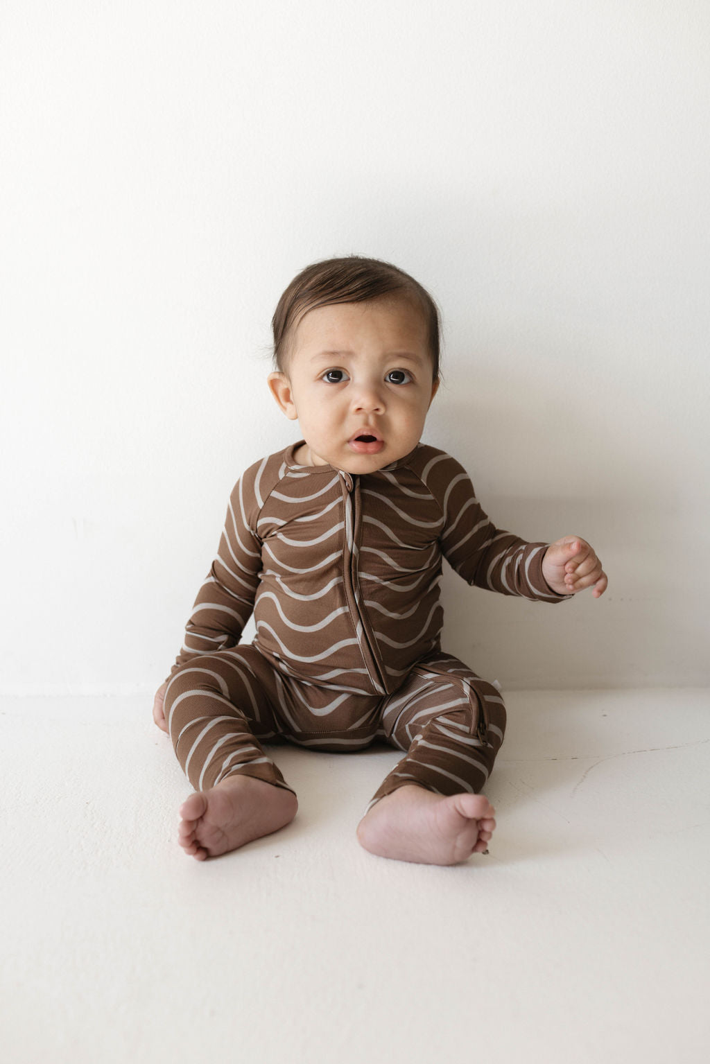 A baby with short dark hair sits against a white wall, dressed in cozy Bamboo Zip Pajamas by forever french baby. The one-piece outfit features light brown and white wavy patterns known as Static Waves, making it perfect for sleepwear. The baby gazes forward with a slightly open mouth, one hand resting on the floor while the other is slightly raised.