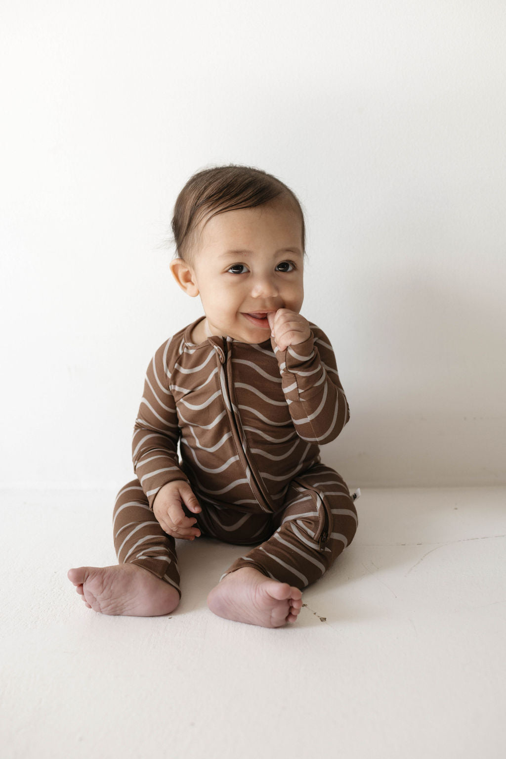 A baby wearing "Bamboo Zip Pajamas | Static Waves" from forever french baby sits on a white surface, smiling and looking slightly to the side while sucking on their thumb. They look cozy in their hypo-allergenic clothing, perfect for sensitive skin.