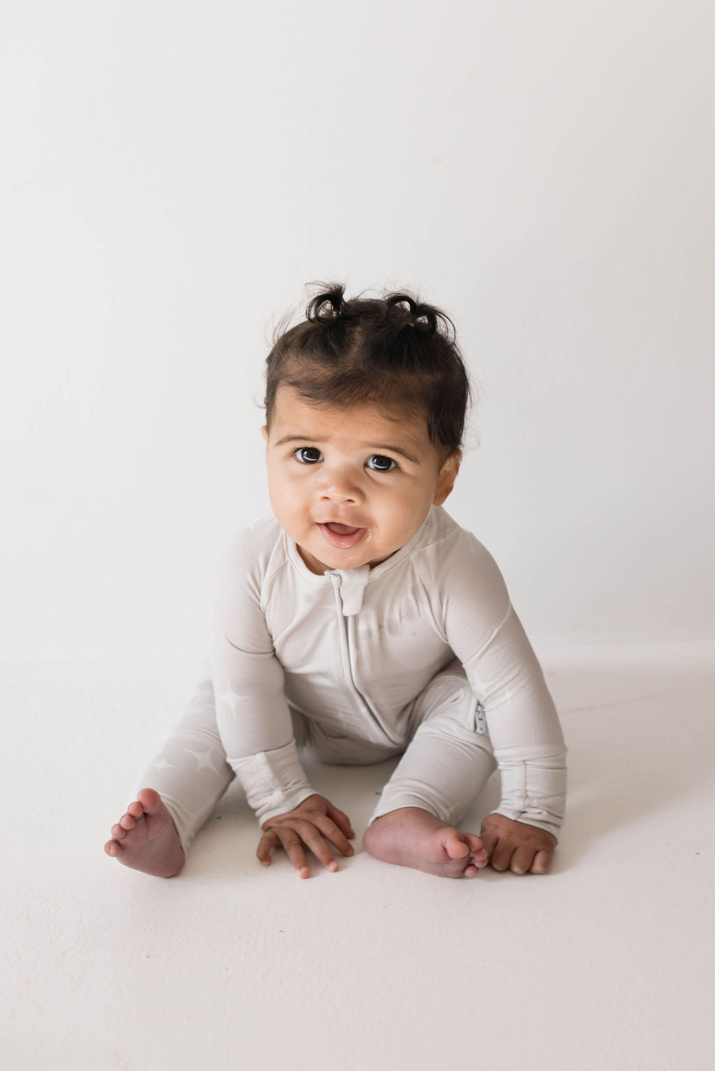 A baby with dark, curly hair sits on the floor wearing a light-colored, long-sleeve onesie made from hypo-allergenic Bamboo Zip Pajamas | Sparkle! | ff club by forever french baby. The baby looks curiously at the camera with wide eyes and slightly parted lips. The background is plain and light-colored.