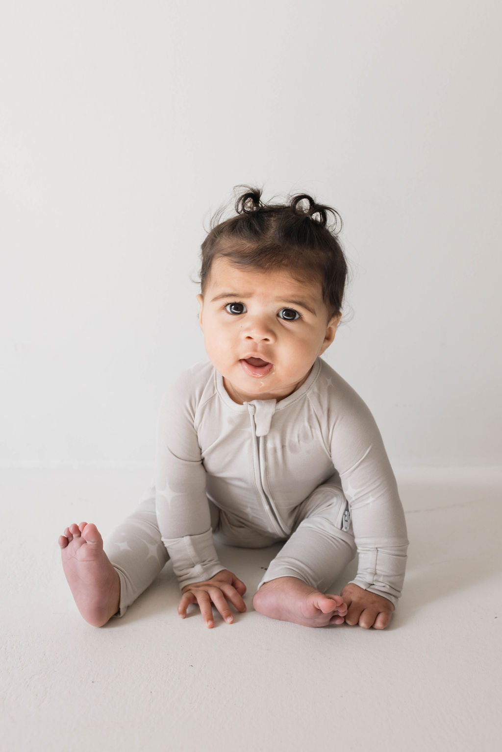 A baby sits on a white surface against a plain white background. The baby, wearing light gray, hypo-allergenic "Bamboo Zip Pajamas | Sparkle! | ff club" from forever french baby, has curly hair styled in small pigtails. With a curious expression and mouth slightly open, the baby looks directly at the camera.