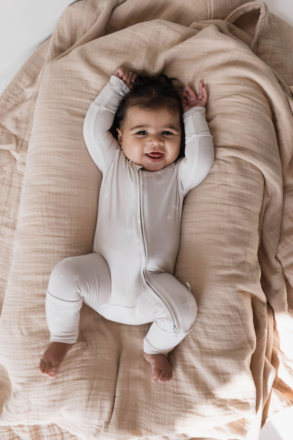 A baby with dark hair lies on a beige blanket, wearing Forever French Baby Bamboo Zip Pajamas in the Sparkle! design—a light-colored, long-sleeved onesie with a zipper. The hypoallergenic clothing keeps the baby comfortable as they raise their arms and smile, looking up.