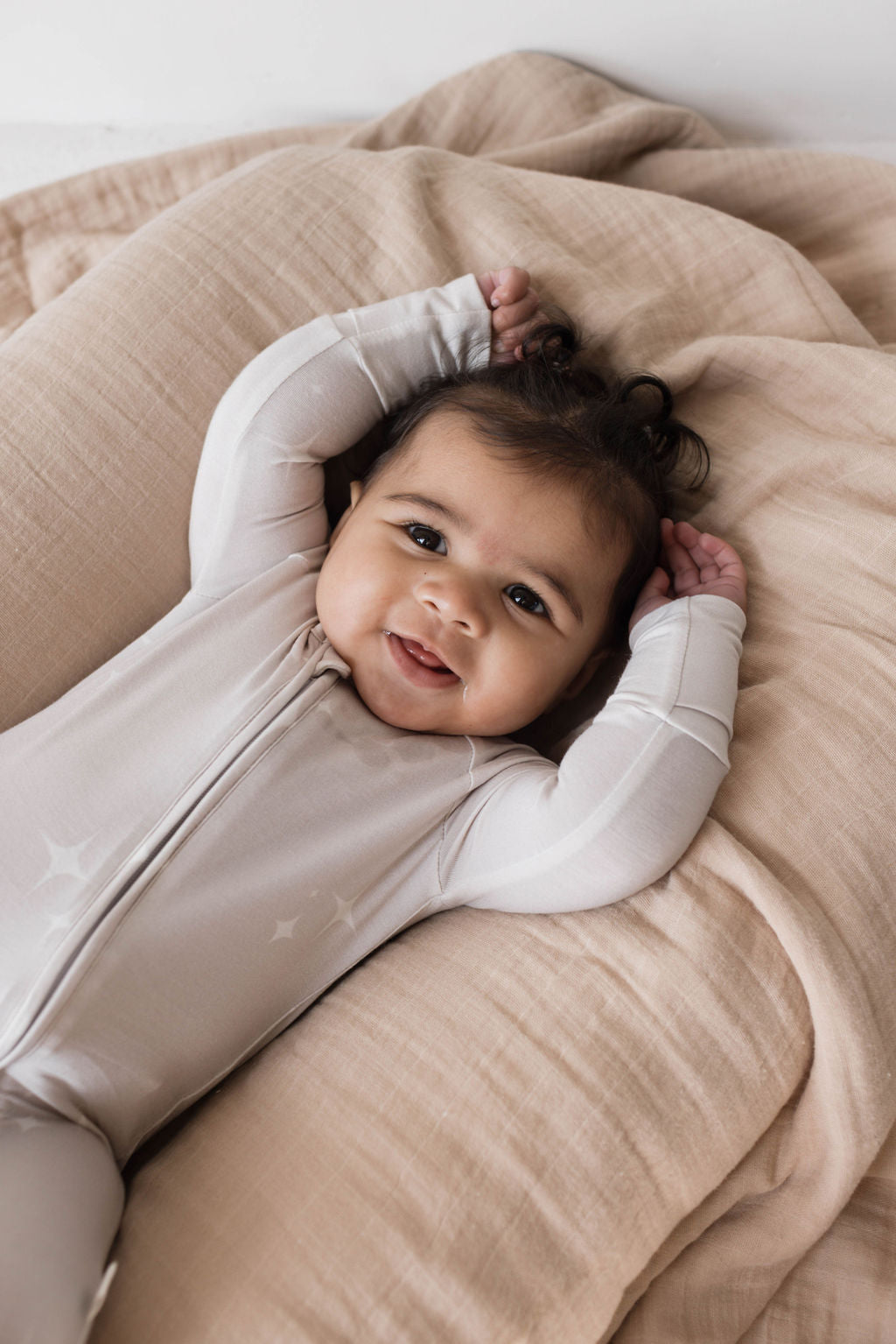 A smiling baby lies on a beige cushioned surface, wearing light-colored Forever French Baby Bamboo Zip Pajamas with a star pattern. The baby's arms are raised above their head, and they are looking up with a joyful expression. Forever French Baby ensures these hypo-allergenic Sparkle! pajamas keep your little one comfortable and happy.