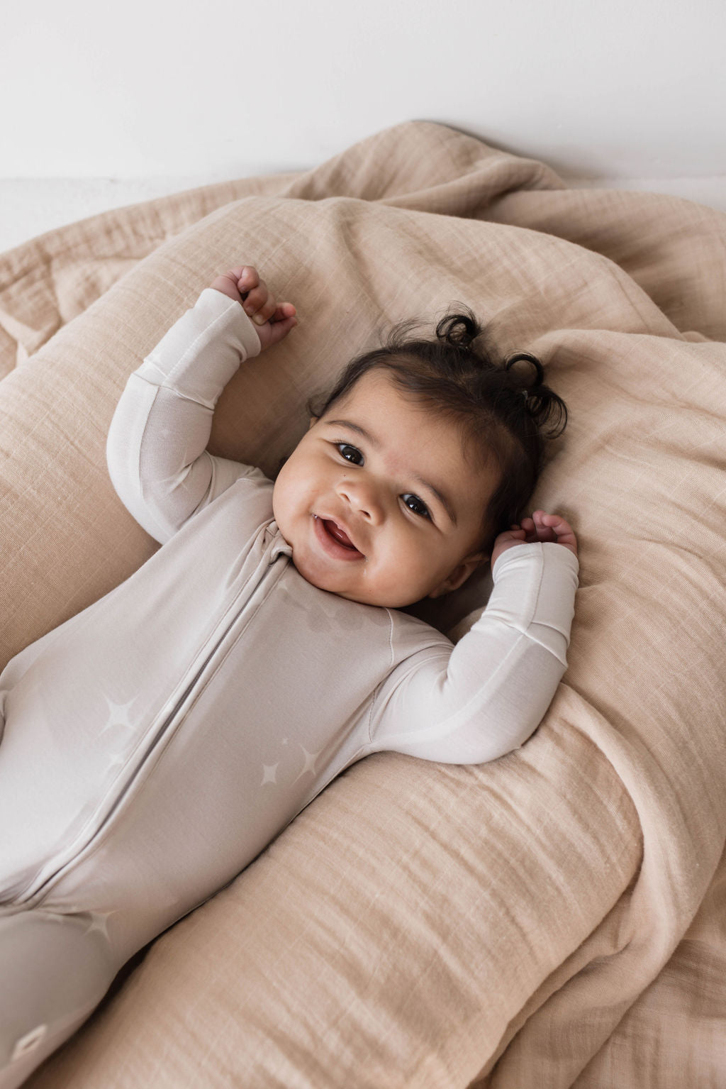 A baby wearing light-colored Forever French Baby 'Bamboo Zip Pajamas in Sparkle!' lies on a plush, beige blanket. The baby is smiling with arms raised, sporting dark hair styled into two small pigtails. The background is soft and neutral-toned, creating a warm and cozy atmosphere.