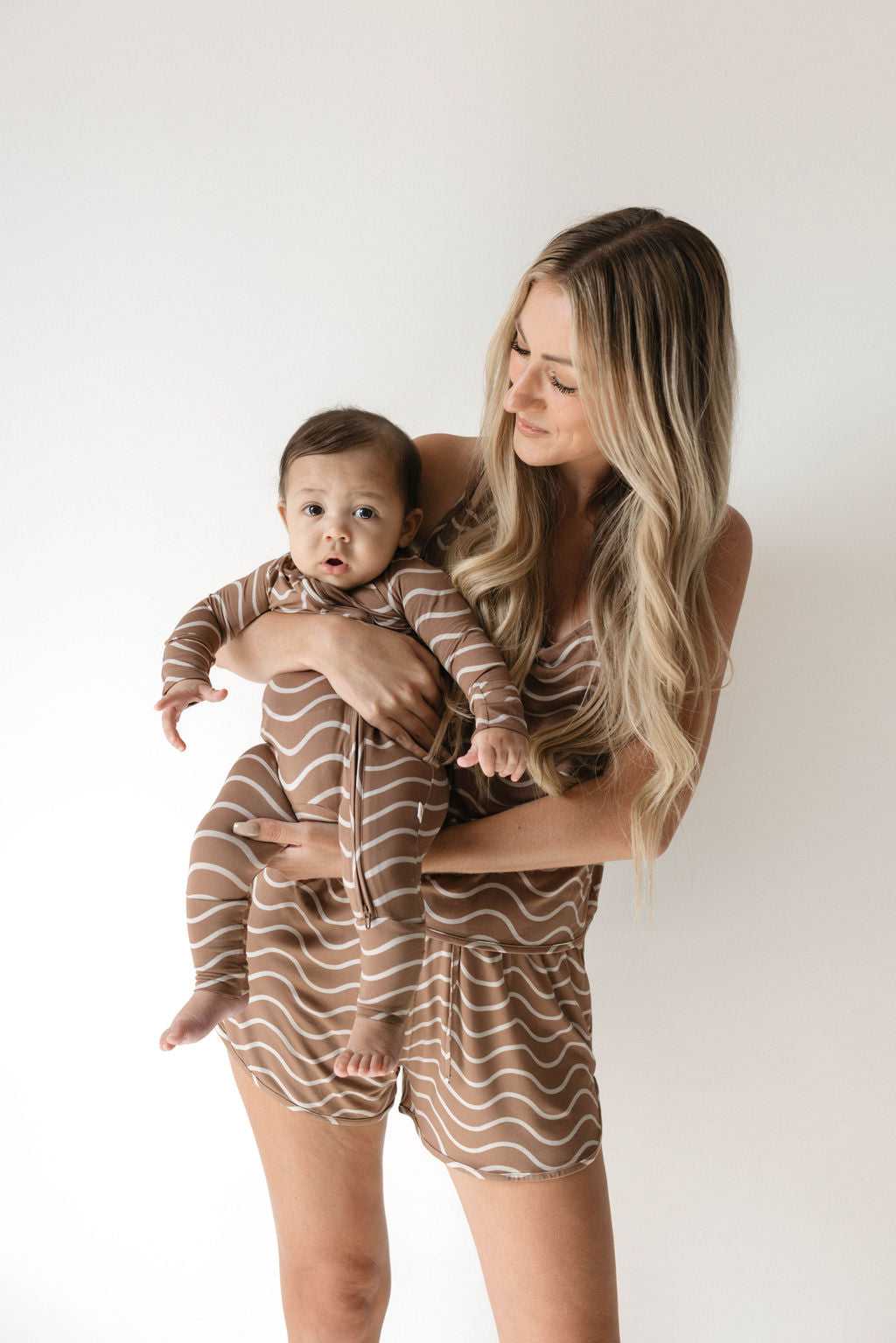 A woman with long blonde hair, wearing a beige outfit, holds a baby dressed in forever french baby's Bamboo Zip Pajamas in the Static Waves pattern, featuring hypo-allergenic beige and white stripes. They are standing against a plain white background. The baby looks at the camera while the woman gazes affectionately at the baby.