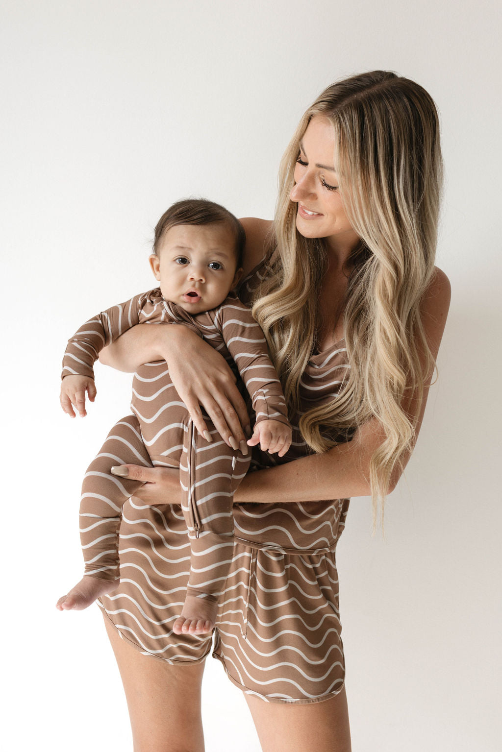 A woman with long blonde hair and a baby are both dressed in matching forever french baby Bamboo Zip Pajamas in the Static Waves pattern. The woman is smiling and looking at the curious baby as they stand against a plain white background.