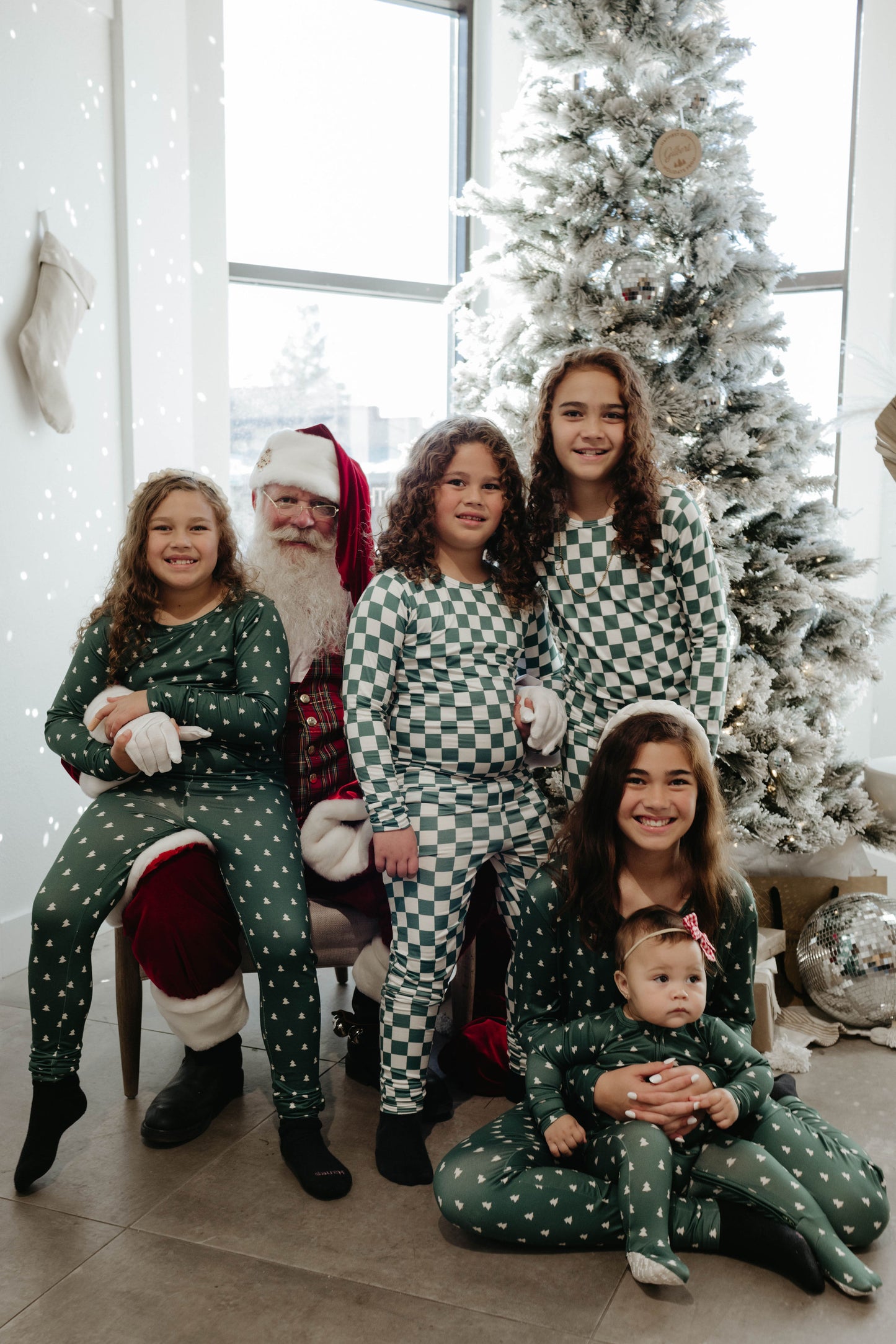 A group of five children in matching green checkered pajamas pose with Santa Claus in front of a decorated Christmas tree. The children are smiling and standing, while the baby, dressed as a Forever French Baby from Amanda Riley Photos' Santa Photos collection, is seated on the lap of an older child who is sitting in front.