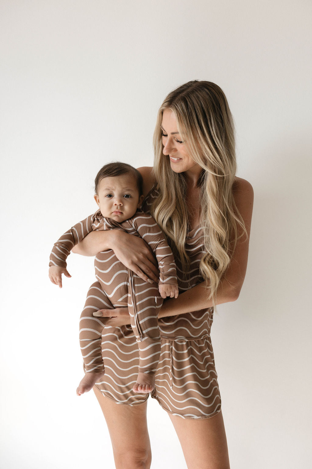 A woman with long blonde hair, dressed in a striped brown and beige outfit, stands holding a baby wearing matching hypo-allergenic Bamboo Zip Pajamas in the Static Waves design from forever french baby. Both are smiling and standing against a plain white background.