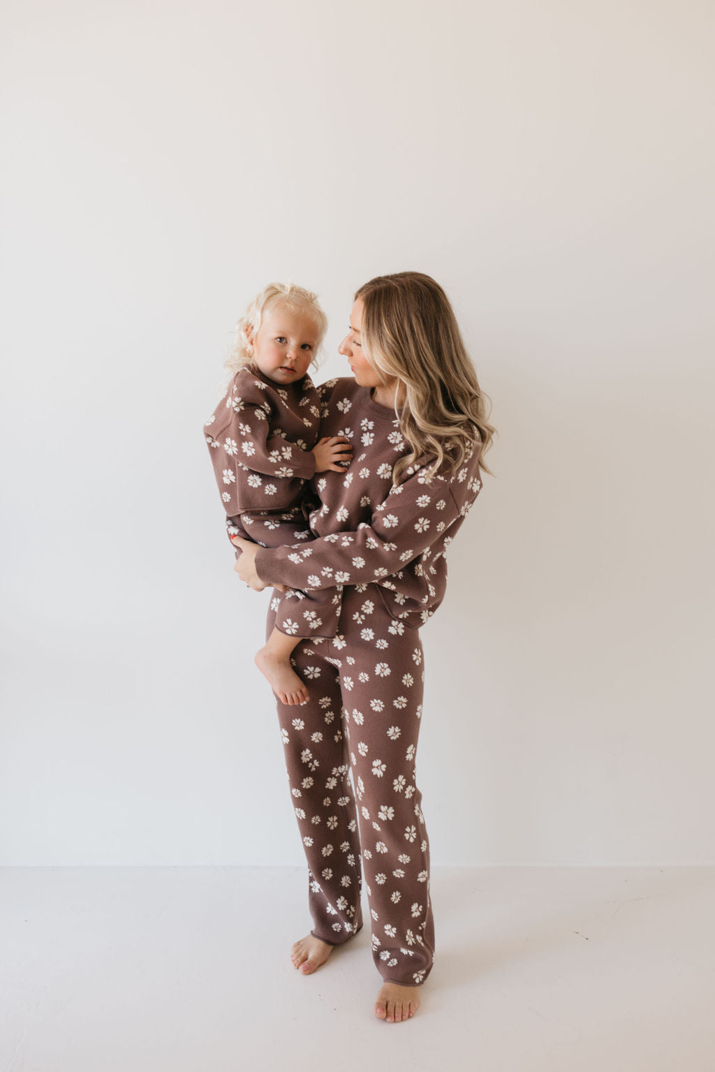 A woman holding a young child, both dressed in the Knit Pant Set | Midnight Meadow by forever french baby, stand barefoot against a plain white background. These matching brown pajamas, adorned with delicate white floral patterns and made from 100% cotton, are perfect for your fall wardrobe, offering both comfort and style.