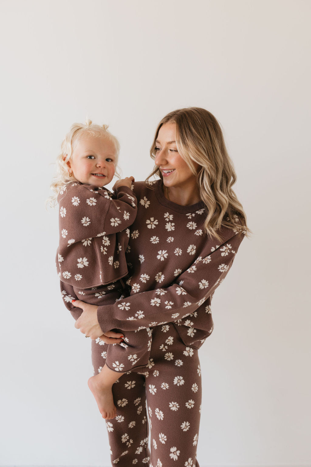 A woman holding a toddler, both clad in the Knit Pant Set | Midnight Meadow by forever french baby, showcase matching knit outfits with brown hues and white floral patterns against a plain off-white background. The woman smiles warmly at the child, who appears happy and relaxed, perfectly capturing cozy vibes ideal for any fall wardrobe.