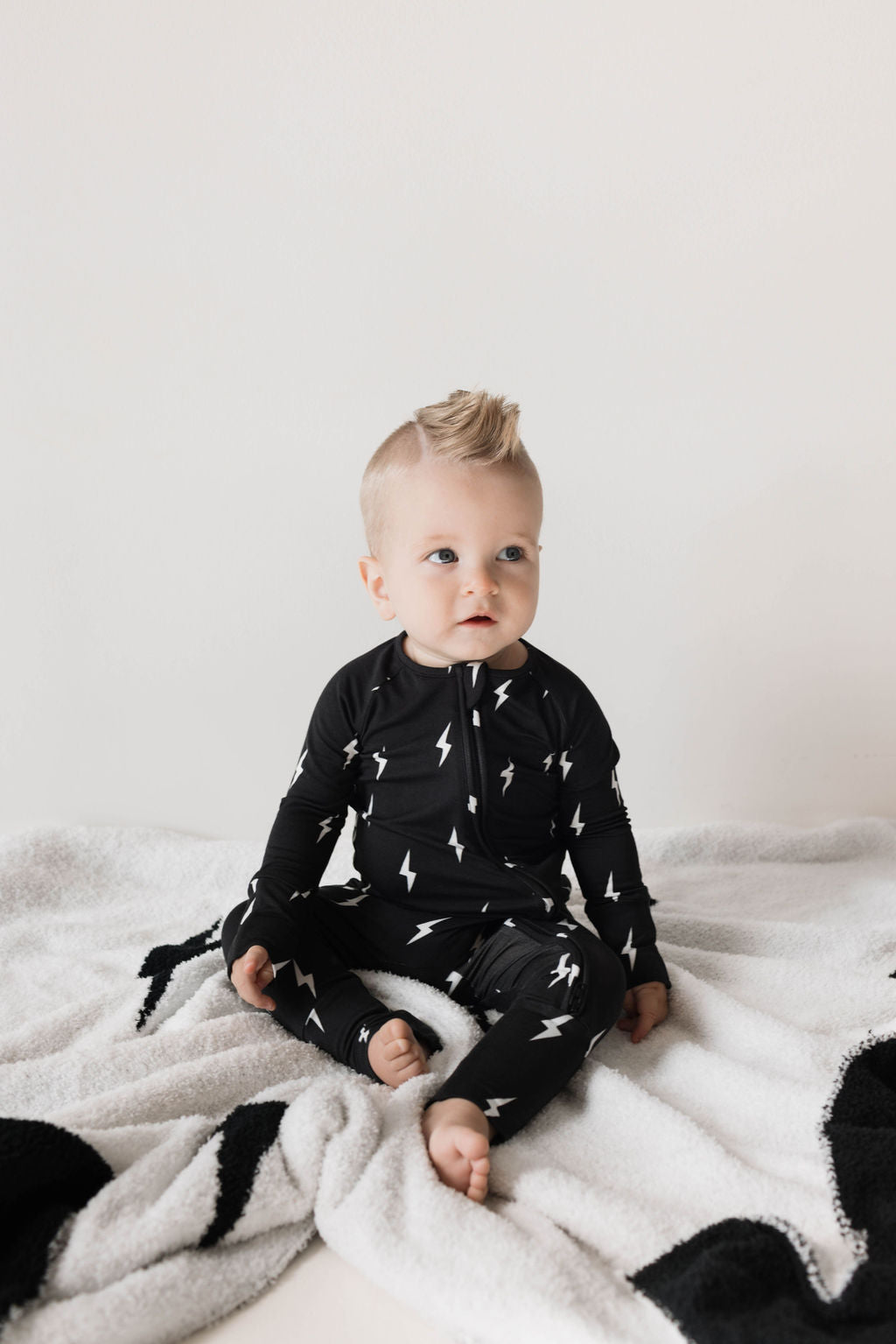 A baby with light-colored hair sits on a soft, white and black patterned blanket. The baby is wearing the Bamboo Zip Pajamas | Midnight Lightning Bolt from forever french baby, decorated with white lightning bolts and gently looks off to the side, nestled in breathable pajamas that ensure comfort.