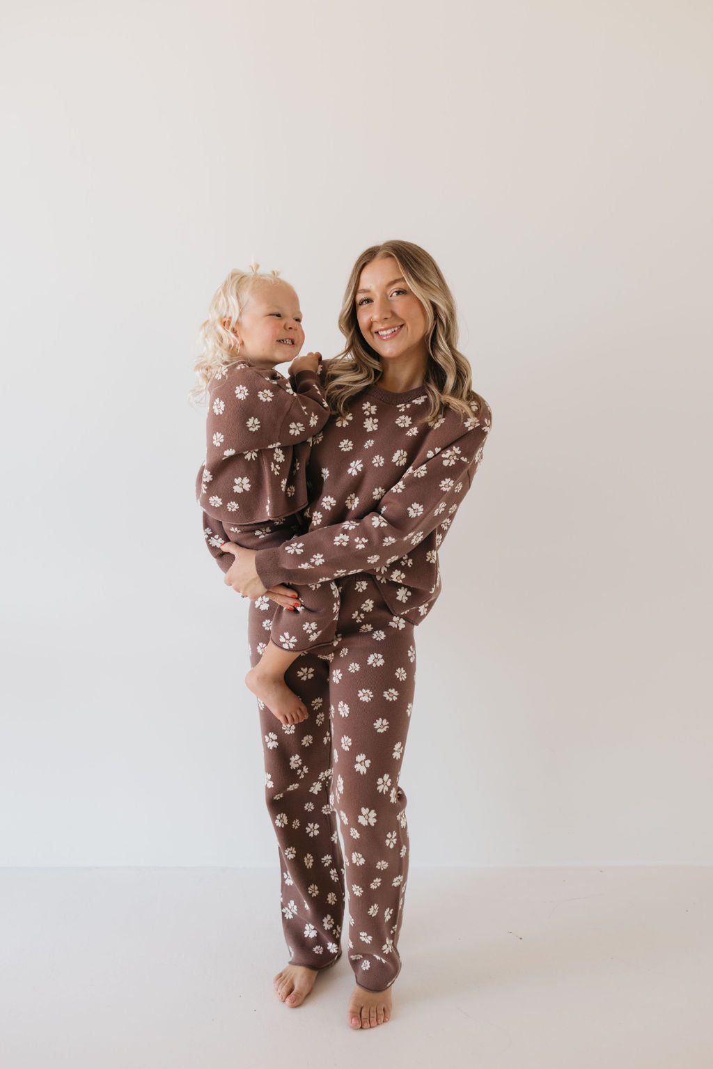 A woman and a young child, both wearing matching outfits from the Knit Pant Set in Midnight Meadow by forever french baby, stand barefoot against a plain white background. The woman holds the smiling child in her arms.