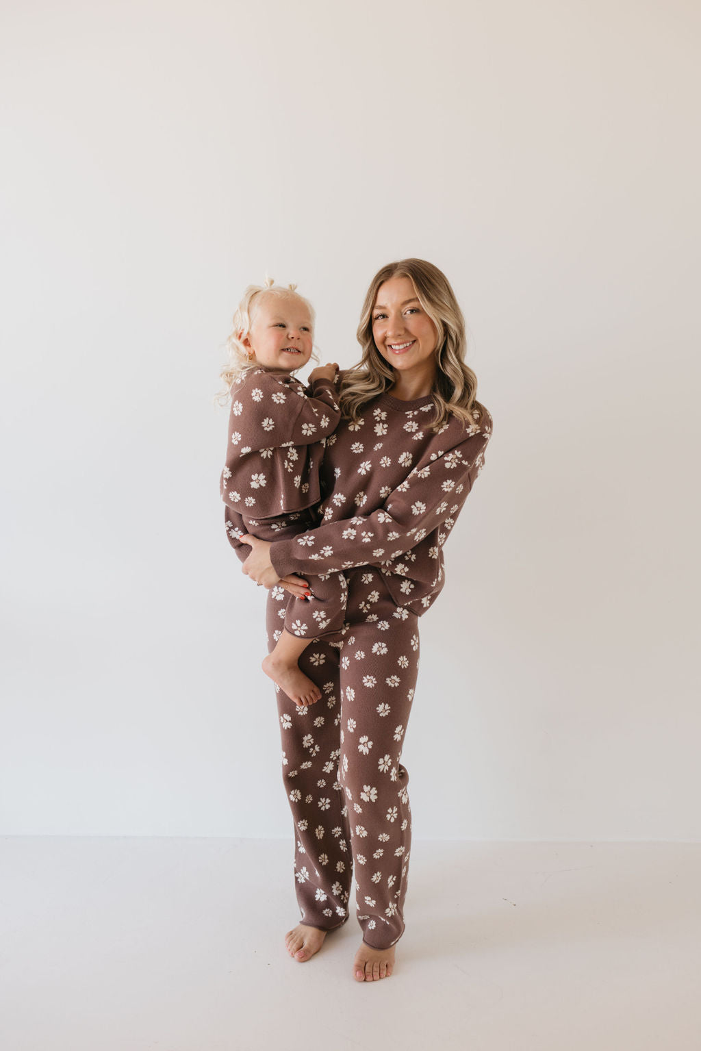 A woman and a child, both with blonde hair, are smiling while wearing matching knit pant sets from forever french baby, featuring the Midnight Meadow design—an elegant touch to their fall wardrobe. The woman is holding the child against a plain white background.