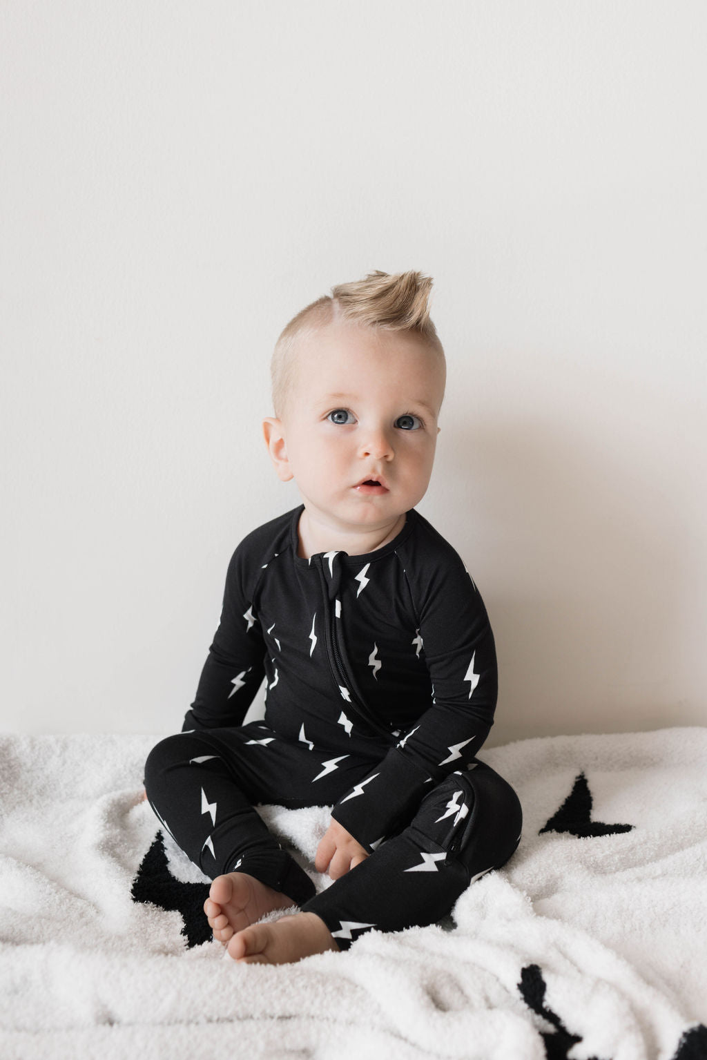 A baby with light skin and light hair sits on a white blanket, wearing hypo-allergenic clothing—a pair of Midnight Lightning Bolt Bamboo Zip Pajamas by forever french baby. The background is a plain white wall, and the baby has a curious expression.