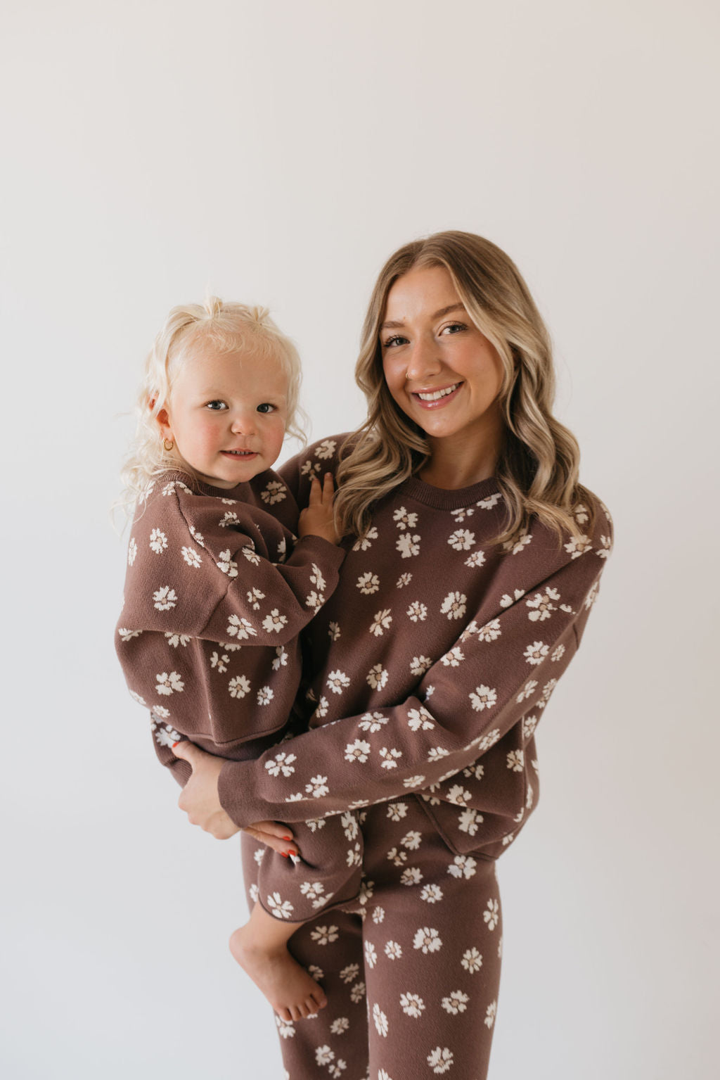 A woman holds a child, both smiling in matching "Midnight Meadow" knit sets from forever french baby, adorned with white flowers, embodying the perfect fall wardrobe against a plain white background.