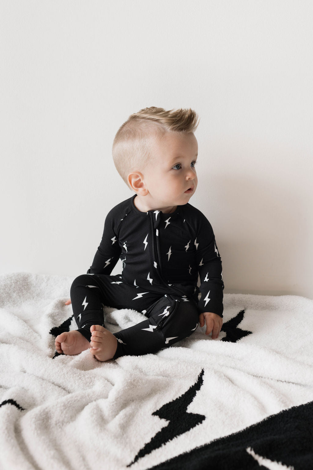 A toddler with short, light hair sits on a white blanket featuring black lightning bolt patterns. The child is wearing the "Bamboo Zip Pajamas | Midnight Lightning Bolt" by forever french baby and looks to the side with an attentive expression, showcasing their breathable pajamas that ensure comfort throughout the day.