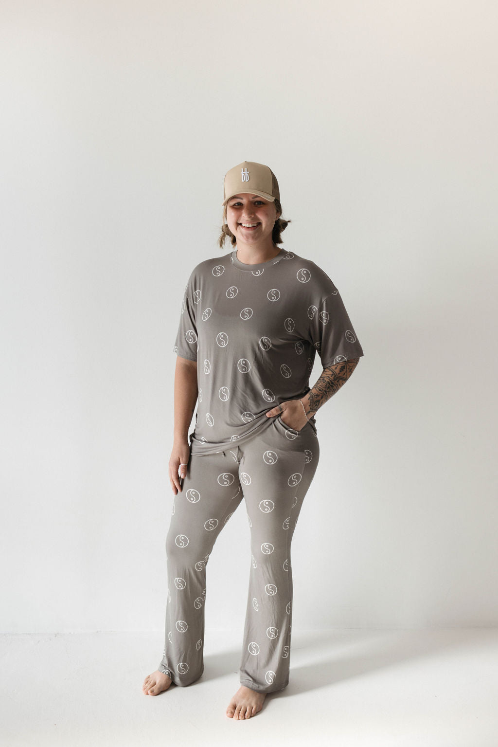 A person stands barefoot against a plain background, smiling and wearing a tan baseball cap and the Short Sleeve Women's Bamboo Pajamas in Sage Yin Yang by forever french baby, which features a matching gray oversized set with white circular patterns. The person has one hand in a pocket and a visible tattoo on their left forearm.