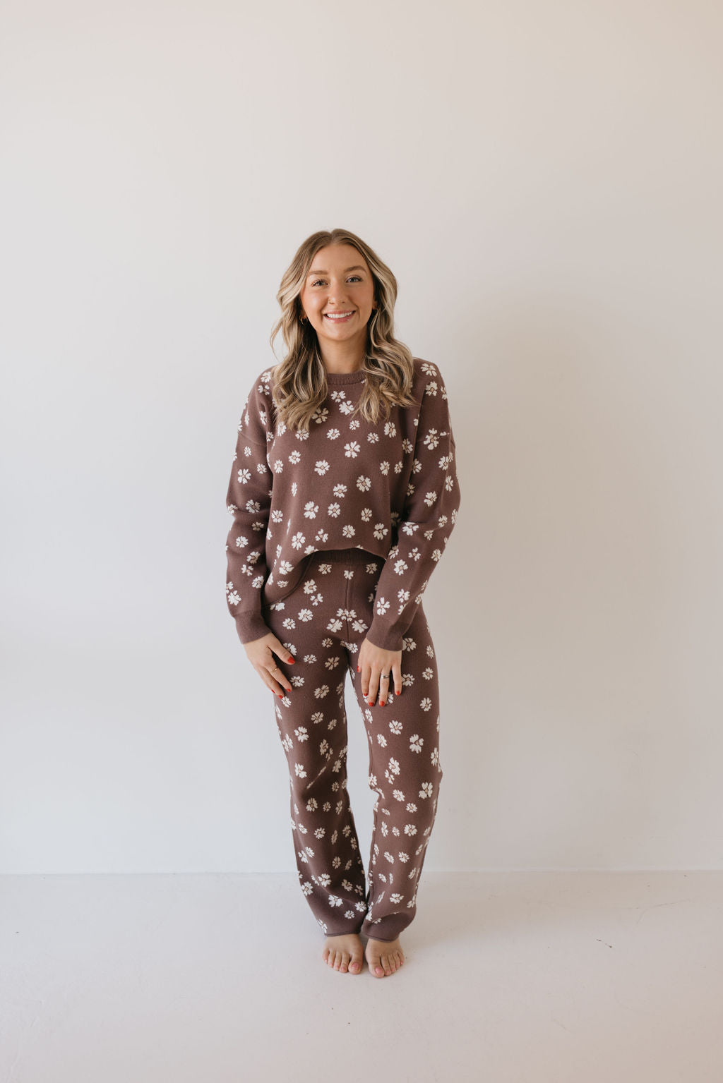 A person stands barefoot against a plain white background, wearing the Women's Knit Pant Set in the Midnight Meadow design from Forever French Baby. The cozy ensemble features a warm brown hue adorned with a delicate white floral pattern, making it an ideal addition to a fall wardrobe. They have long, wavy hair and are smiling at the camera.