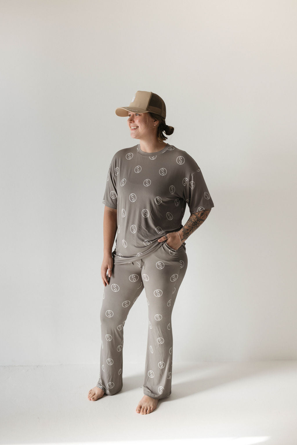 A person with long hair stands barefoot on a white floor, wearing an oversized Short Sleeve Women's Bamboo Pajamas in Sage Yin Yang from forever french baby. They also wear a tan baseball cap and have tattoos on one arm. Their hands are partly in their pockets, and they smile while looking to the side.