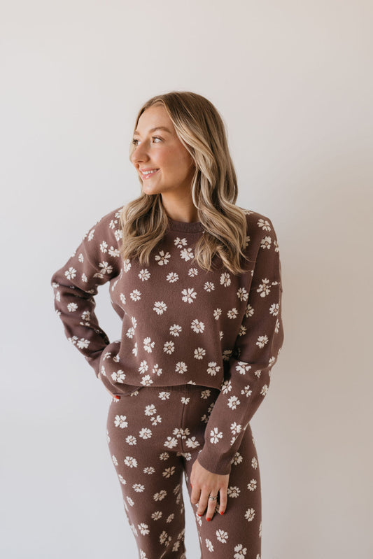 A person with long hair smiles while looking to the side, wearing the Women's Knit Pant Set from forever french baby in the Midnight Meadow design, featuring a brown knit adorned with white floral patterns. They stand against a plain light background, embodying the cozy essence of a fall wardrobe.