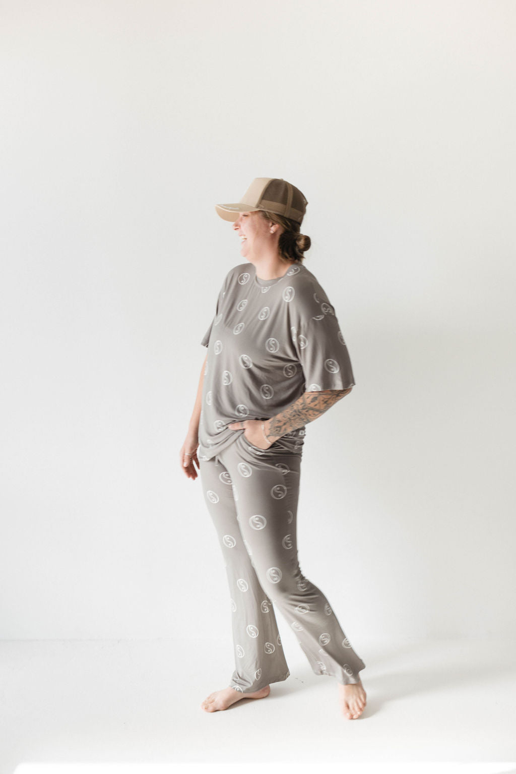 A person wearing the "Short Sleeve Women's Bamboo Pajamas | Sage Yin Yang" by forever french baby, featuring an oversized fit with circular patterns, consisting of a short-sleeved top and pants. They are barefoot, looking to the side, and wearing a beige cap against a plain white background.