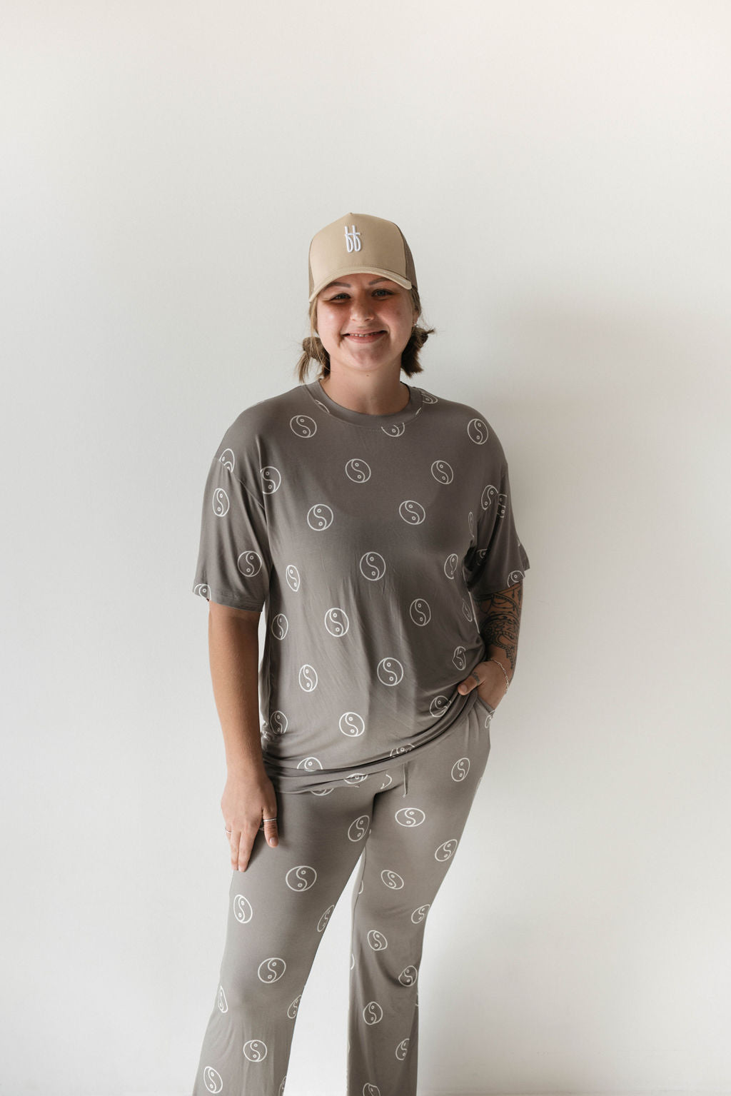 A person stands against a plain white background, smiling, in a matching set of Short Sleeve Women's Bamboo Pajamas in Sage Yin Yang by forever french baby. The pajama set, made from breathable fabric and featuring a circular logo pattern, is complemented by a beige baseball cap with a white logo. They have one hand casually tucked into their pocket.