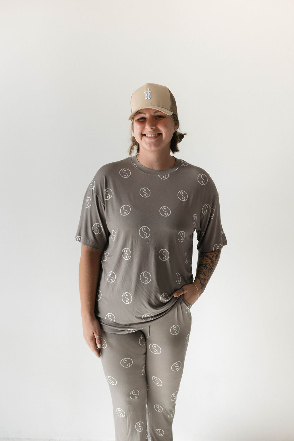 A person stands against a white background, wearing a matching beige outfit featuring the Sage Yin Yang circular logo pattern from forever french baby. They also sport a beige baseball cap. The breathable ensemble, reminiscent of an oversized pajama set, includes one hand casually in their pocket as they smile at the camera.