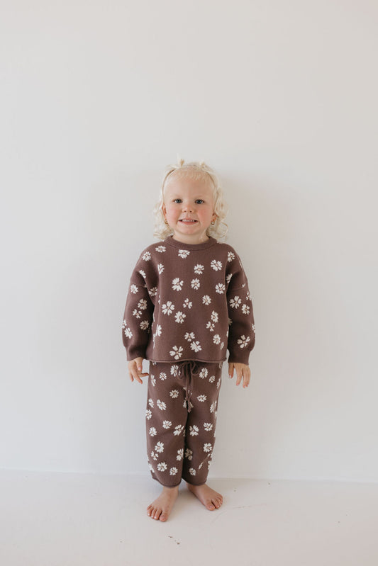 A young child with curly blond hair is standing barefoot against a plain white background, wearing the Knit Pant Set | Midnight Meadow from forever french baby. The matching brown knit set features a long-sleeved top and pants adorned with delicate white floral patterns, crafted from 100% cotton. The child smiles at the camera, ready for their fall wardrobe debut.