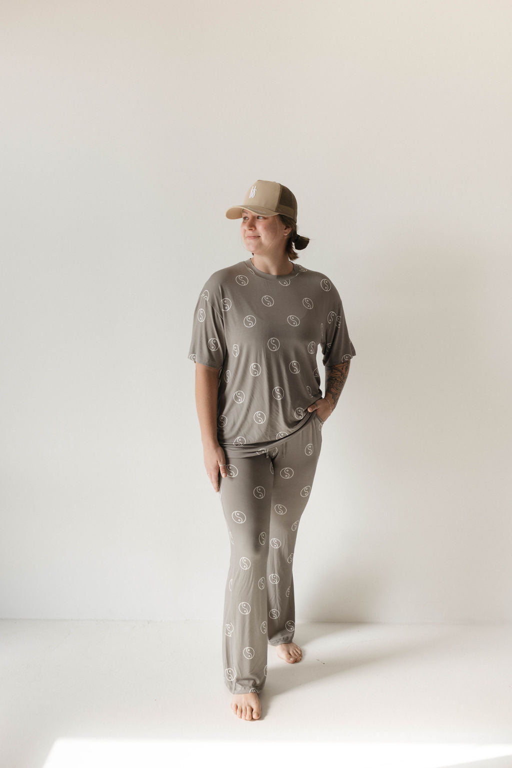 A person wearing a taupe baseball cap and a matching Sage Yin Yang Short Sleeve Women's Bamboo Pajama set from forever french baby stands barefoot on a light-colored floor. Their breathable outfit, reminiscent of an oversized pajama set with white circular patterns, features one hand in their pocket as they look off to the side with a neutral expression.