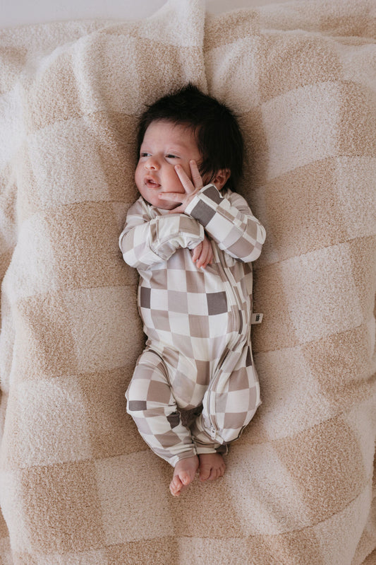 A baby clad in Forever French Baby's Bamboo Zip Pajamas, featuring the Into the Woods Checkerboard pattern, lies on a soft, patterned blanket. The baby's eyes are open, with one hand resting on their cheek. The beige and white bamboo pajamas provide breathable comfort ideal for delicate skin.