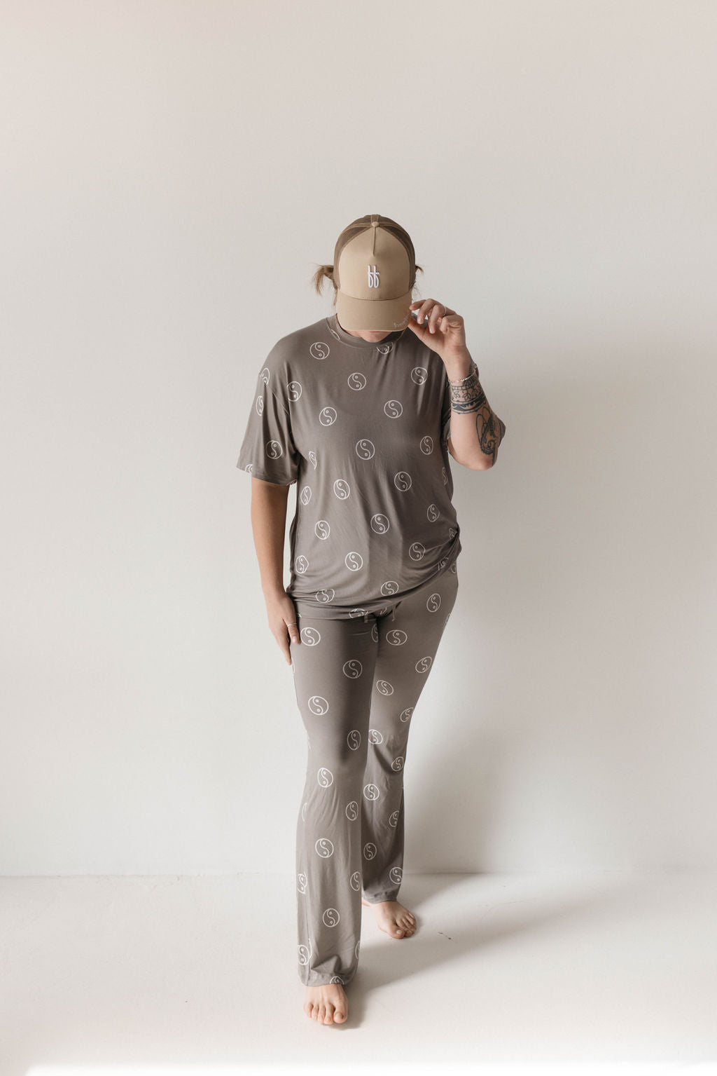 A person stands barefoot against a plain white background, wearing an oversized pajama set called the "Short Sleeve Women's Bamboo Pajamas | Sage Yin Yang" by forever french baby. The breathable clothing, in a subtle gray pattern, consists of a short-sleeved shirt and pants. Adding to the ensemble is a beige baseball cap, slightly tilted forward, partially obscuring their face.