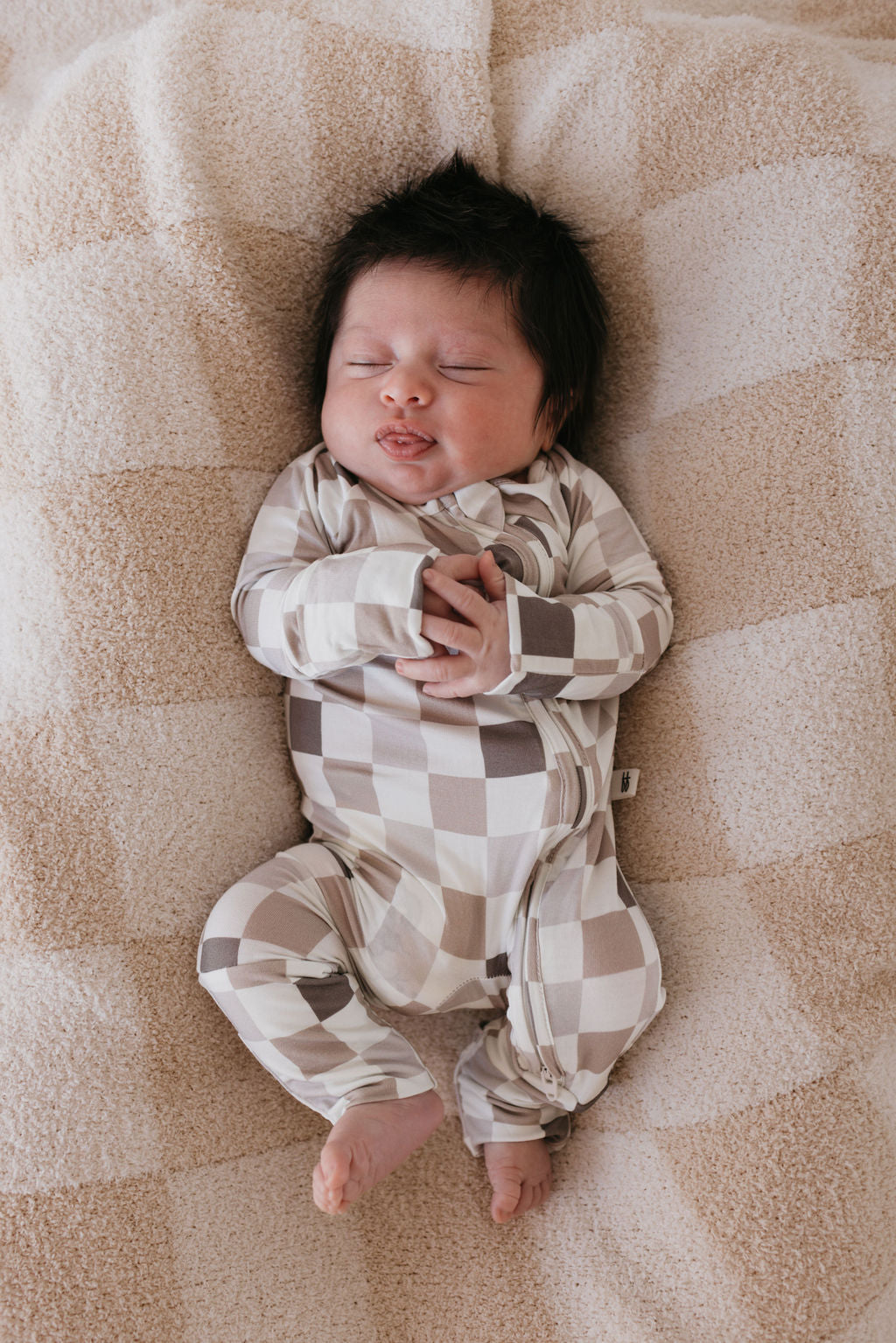 A sleeping baby dressed in forever french baby's Bamboo Zip Pajamas | Into the Woods Checkerboard lies on a soft, patterned blanket. The hypo-allergenic fabric ensures comfort as the baby with dark hair rests peacefully, hands gently clasped. The cozy setting conveys warmth and serenity.