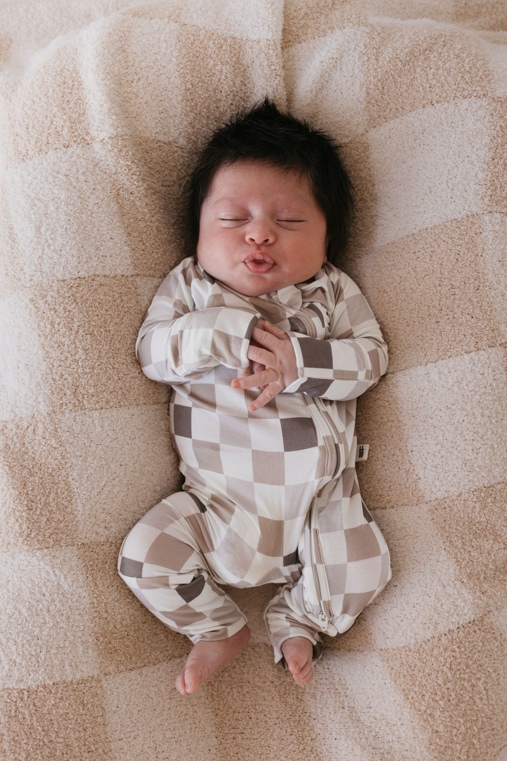 A sleeping baby lies on a soft, beige checkered blanket. Dressed in the hypo-allergenic, breathable Bamboo Zip Pajamas from forever french baby's Into the Woods Checkerboard collection, the baby rests peacefully with eyes closed and lips pursed.