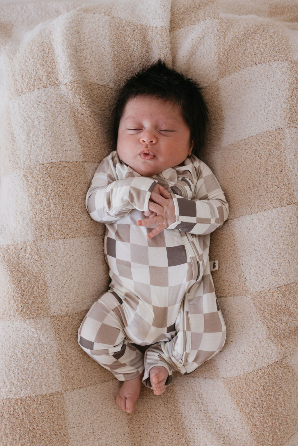 A baby with dark hair sleeps peacefully on a soft, checkered blanket. The baby is clad in hypo-allergenic Bamboo Zip Pajamas in the "Into the Woods Checkerboard" pattern by forever french baby, hands clasped near the chest, lying on a cozy textured surface.