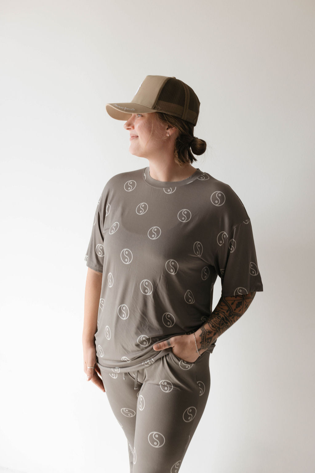 A person with a tattoo on their right forearm stands against a plain light background, wearing breathable Short Sleeve Women's Bamboo Pajamas by forever french baby in the Sage Yin Yang pattern and a tan baseball cap. They have their left hand in their pocket and are looking to the side.