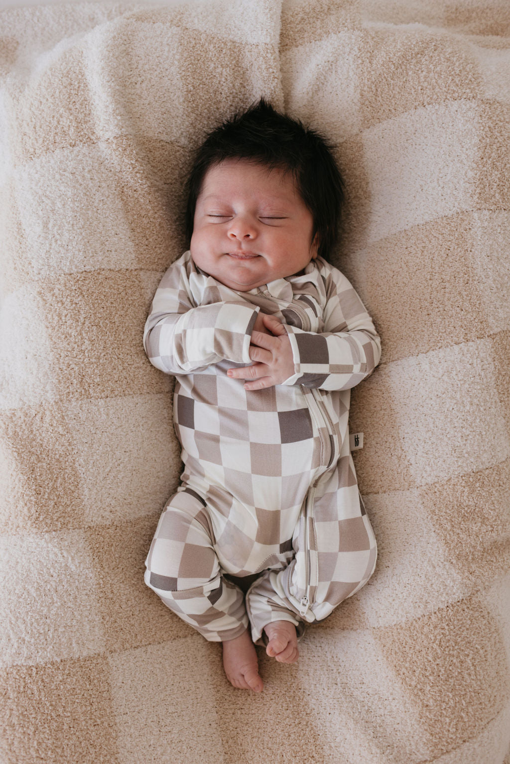 A sleeping baby in a cozy, forever french baby Bamboo Zip Pajamas featuring the Into the Woods Checkerboard pattern, made from breathable, hypo-allergenic bamboo material, lies on a soft, beige blanket. The dark-haired baby is peacefully resting with hands clasped over their chest, creating an image that exudes warmth and tranquility.