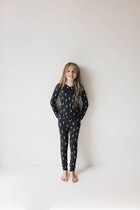 A child with long hair stands barefoot against a white background, wearing matching dark Bamboo Two Piece Pajamas | Midnight Lightning Bolt from forever french baby. They are smiling and have their hands in the pockets of their pajama pants, which are decorated with a pattern of white lightning bolts.