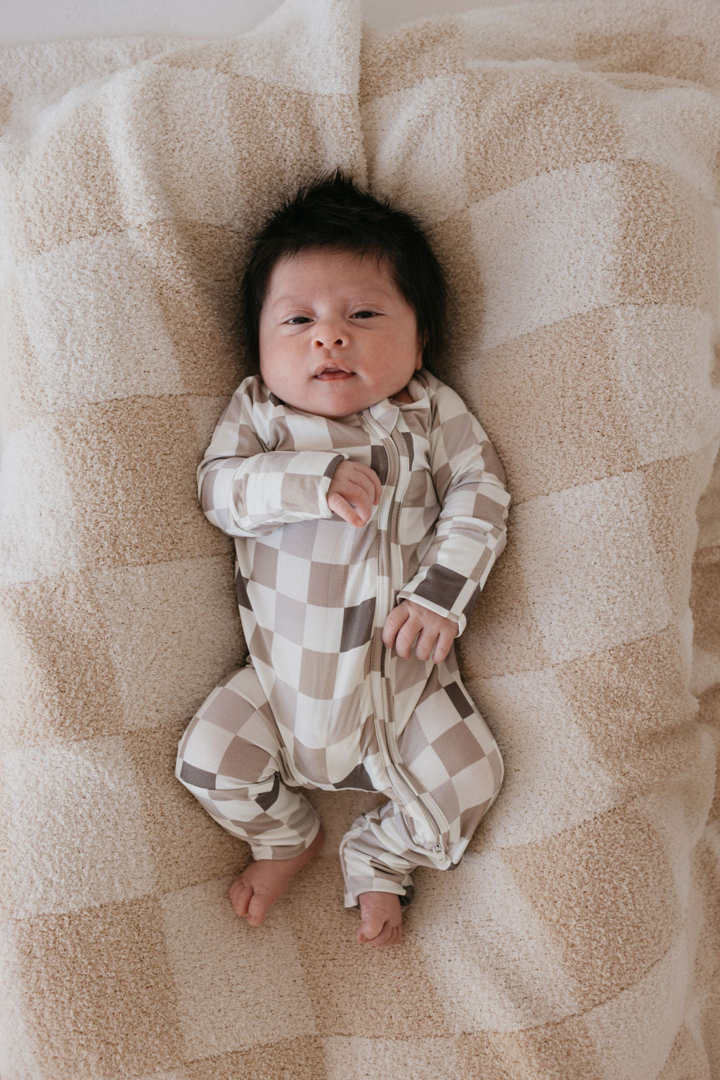 A baby with dark hair is lying on a plaid-patterned blanket, snuggled in the "Into the Woods Checkerboard" Bamboo Zip Pajamas by forever french baby. The baby gazes up with a curious expression, hands near their face, and toes pointing outward, enjoying the softness of breathable hypo-allergenic bamboo fabric.