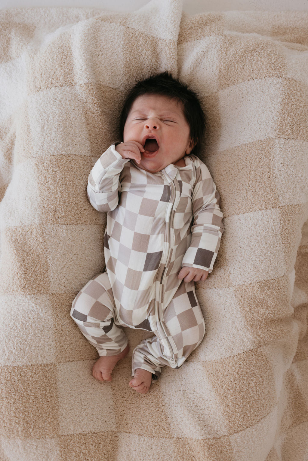 A baby wearing the hypo-allergenic Bamboo Zip Pajamas in Into the Woods Checkerboard pattern by forever french baby is lying on a soft, checkered blanket. The baby is yawning with eyes closed and one hand near their face.