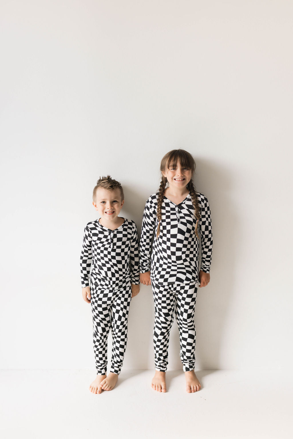 Two children stand against a white background, both dressed in matching Bamboo Two Piece Pajamas in Black & White Wavy Checkerboard by forever french baby. The child on the left is shorter with short hair, while the child on the right is taller with long hair styled in braids. Both kids are barefoot and smiling.