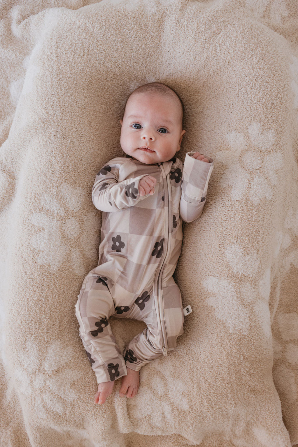 A baby wearing the Bamboo Zip Pajamas in the Flower Child design from forever french baby lies on a soft, fluffy blanket. The beige and brown checkered onesie adorned with flower patterns envelops the baby, who looks up with an inquisitive expression while resting comfortably.